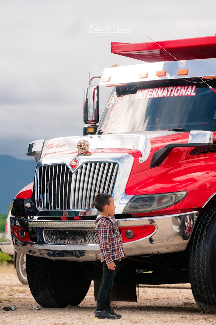 A young boy stands captivated by a large red semi-truck parked outdoors, showcasing vibrant contrasts.
